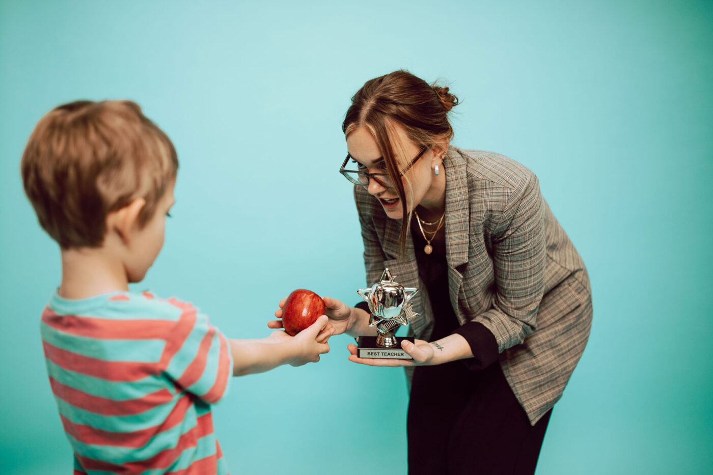 Kind geeft een rode appel aan een juf die een trofee met ‘Best Teacher’ vasthoudt.