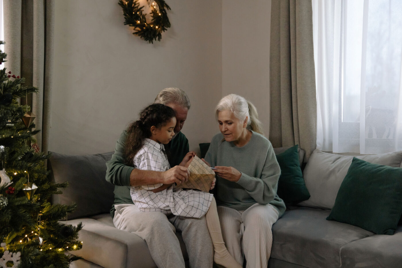 Een meisje zit met een oudere man en vrouw op de bank bij een ingepakt cadeau en een kerstboom.